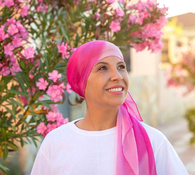 A woman wears a pink headscarf and stands in front of a tree blooming with pink flowers.